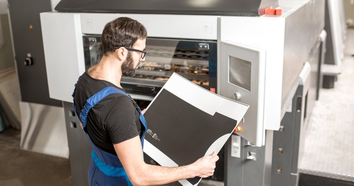 Home » Latest Printing News A worker in a print shop inspects a large, printed sheet in front of a professional offset printing press.