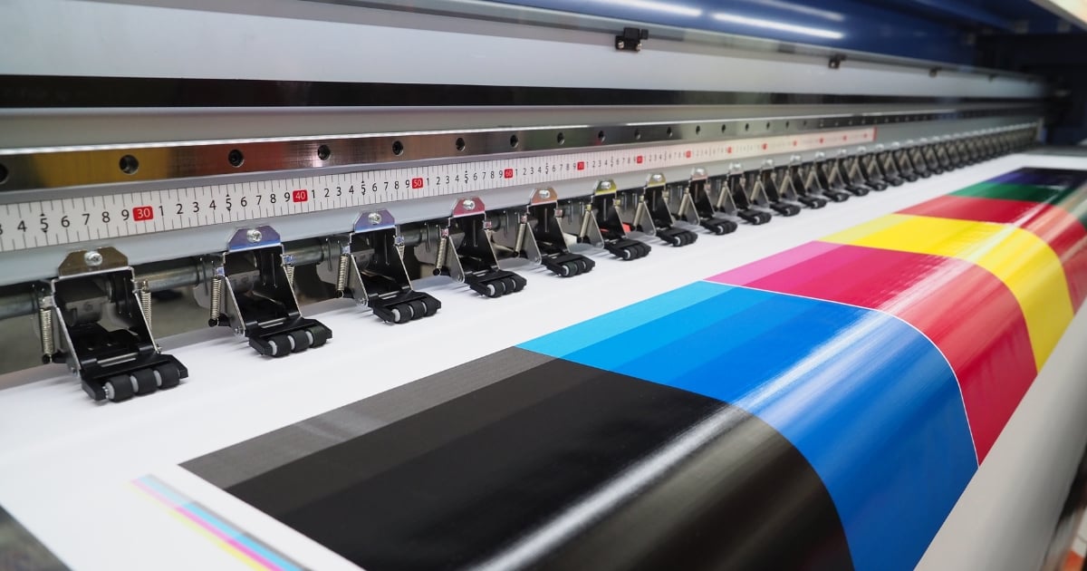 Home » Latest Printing News A wide-format industrial inkjet printer outputting a large vinyl sheet with vibrant blocks of CMYK colours and greyscale test patterns.