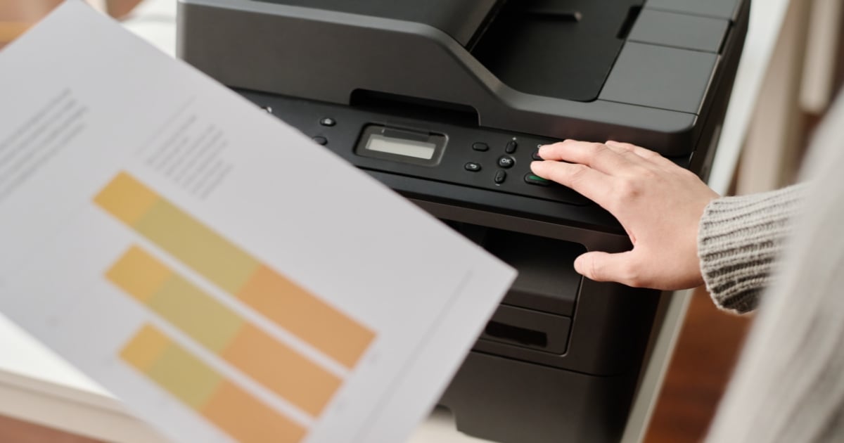 Home » Latest Printing News A person pressing a button on a black multi-function office printer while holding a document featuring yellow and orange bar charts.