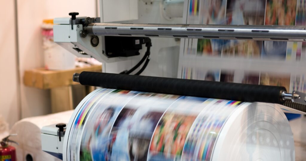 A large industrial printing press rapidly processing a continuous roll of glossy paper printed with colourful photographic images and CMYK colour bars.